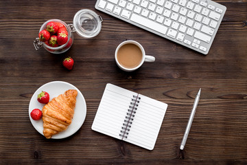 Light summer lunch at workplace. Coffee, strawberry, croissant near keyboard on wooden background top view copyspace