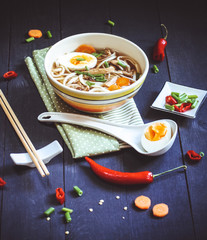 Chicken soup with noodles in a plate on a dark wooden background with hot red pepper carrots and onions. Egg in a white spoon and asian sticks.