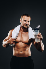 A handsome young sportsman standing half naked with a towel in his neck , Bodybuilder after a workout with a shaker on  dark background