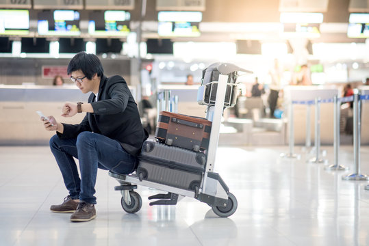 Young Asian man checking time sitting on airport trolley with his suitcase luggage in the international airport terminal, business travel and flight delayed concepts