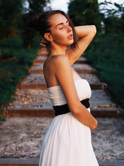 Naklejka premium Young brunette model at the white dress staying at the stairs with green trees with closed eyes