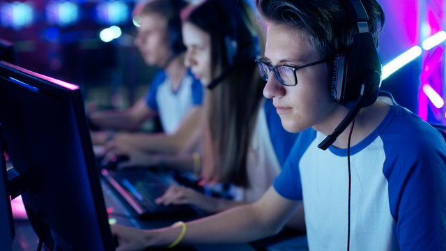 Professional Boy Gamer Plays in Video Game on a eSports Tournament/ Internet Cafe. He Wears Glasses and Headphones with Microphone. Other Girls and Boys Players Playing in Background.