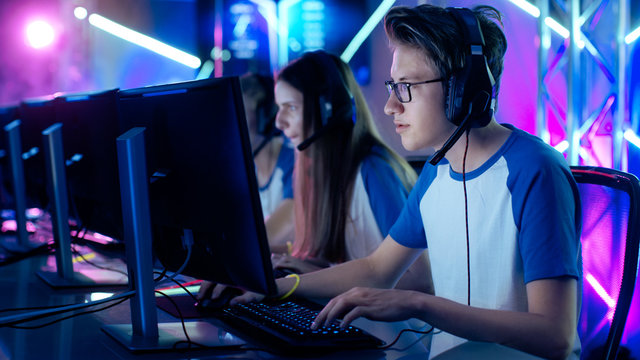 Team of Professional Cybersport Gamers Playing Video Games on a Cyber Games Tournament. Girls and Boys Have Headphones On, Arena is Lit with Neon Lights.