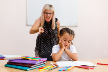 Angry teacher yelling. the teacher screams at the little schoolgirl. little girl with closed eyes sits at a desk in the classroom