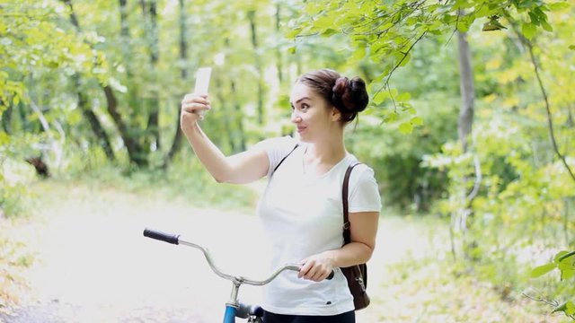 An amusing girl with a backpack and a bicycle makes selfie photo on her phone.
