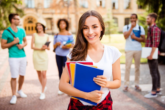 Focused shot of cheerful smart brunette caucasian girl is holding note books, smiling, standing near college building, her friends are behind, they passed tests, so cheerful and carefree!