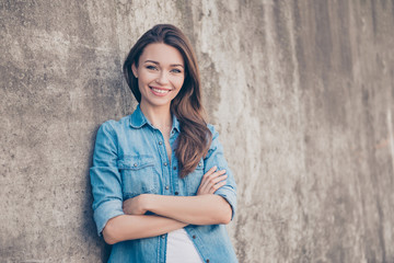 Adorable young brunette lady is leaning the concrete wall outdoors, smiling, with crossed arms, in casual denim jeans outfit