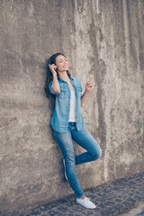 Fototapeta na wymiar Enjoy the music! Young cute hipster is leaning the concrete wall in the city, listening to the music, gesturing, having fun, in modern denim outfit