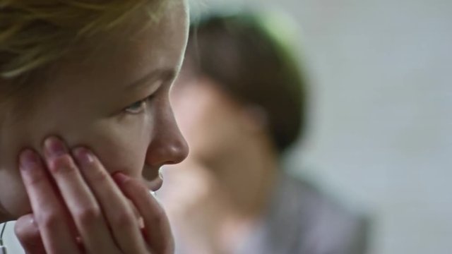 PAN with close up of sad teenage girl with short blond hair listening to music in earphones and looking away as her worried mother talking to female psychiatrist