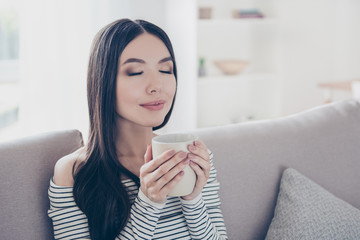 Good morning! Close up portrait of charming dreamy korean young lady drinking hot tea. She is relaxed, in casual wear, sitting on the couch, with closed eyes