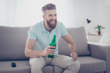 Young happy smiling bearded man in casual clothes sitting on sofa with bottle of beer and watching sport games on tv