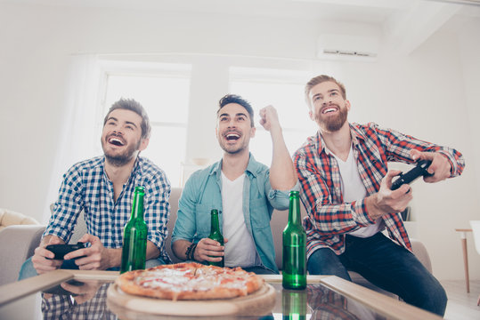 Yes! Team of winners! Bachelor men`s life. Low angle of three happy joyful men, sitting on sofa and playing video games with beer and pizza, smiling, gesturing, enjoying themselves
