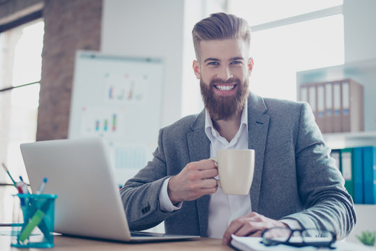 Happy smiling bearded man in jacket drinking coffee in the morning before work, sitting at the light modern office, he has a beaming smile