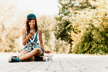 Attractive urban girl sitting on skateboard and  using a mobile phone.