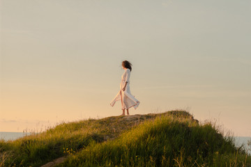 young woman walking on the morning beach in beautiful white dress. Fit female having good time during turing the sunrise. 
