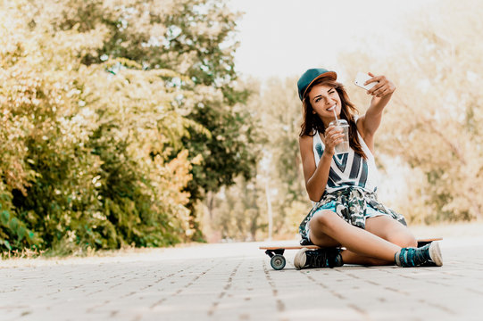 Cute brunette girl taking photo with smartphone while sitting on longboard.