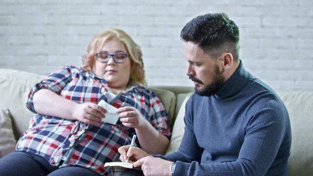 PAN of obese woman with handkerchief sitting on sofa and talking to male psychotherapist making notes