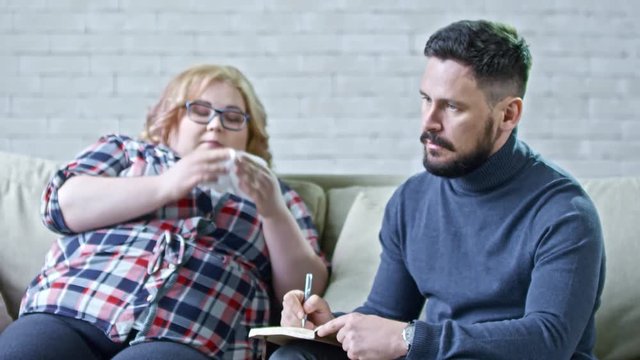 Medium shot of serious male psychotherapist sitting on sofa beside crying obese woman and making notes