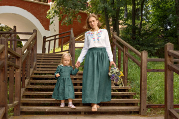  Family mom with daughter in vintage retro style linen dresses  with bouquet of flowers walking dawn wooden stairs in a park garden