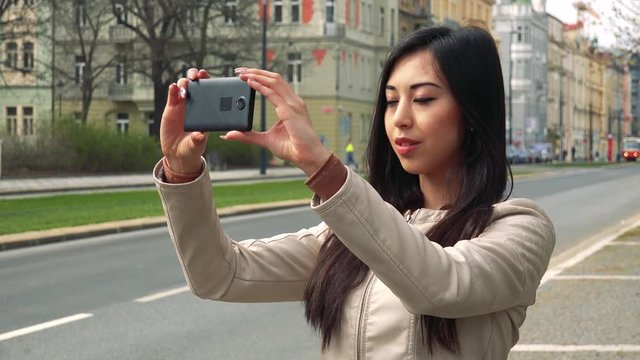 A young Asian woman takes pictures of her surroundings with a smartphone in a street in an urban area