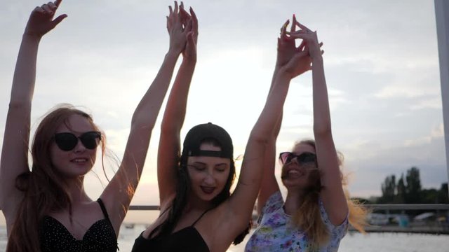 happy best girlfriends dancing in the sunset on beach, three female raises arms up on seaside, summer evening, young women dance on background shiny sea in vacation, girls have fun on embankment