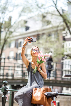 Young woman making selfie photo with bouquet of pink tulips sitting on the fence near the water channel in Amsterdam city