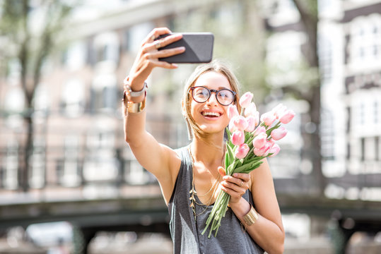 Young woman making selfie photo with bouquet of pink tulips sitting on the fence near the water channel in Amsterdam city