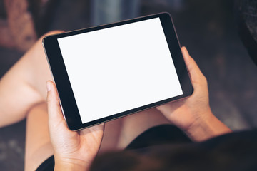 Mockup image of woman's hands holding black tablet pc with blank white screen on thigh with concrete floor background in modern cafe