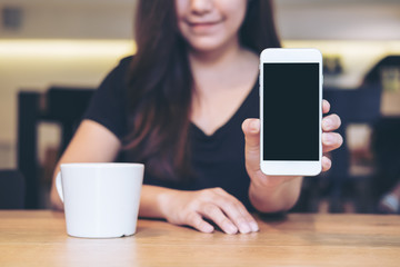 Mockup image of an Asian beautiful woman holding and showing white mobile phone with blank black screen and white coffee coupon wooden table in cafe