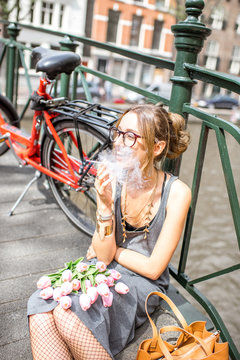 Young beautiful woman smoking a cigarette sitting with flowers and bicycle on the bridge in Amsterdam city