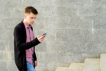 Stylish young man in plaid shirt and jeans walking up stairs and using smartphone outdoors. Student with cellphone walking up stairs, free space