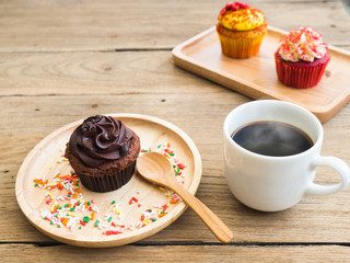 chocolate cupcake put on a spherical wooden plate. Beside of cupcake have Vintage alarm clock and white coffee mug.In the background have yellow cupcake and Red cupcakes.