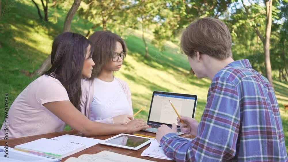 Poster Multiethnic group of concentrated young students sitting and talking outdoors while using laptop computer. Looking aside.