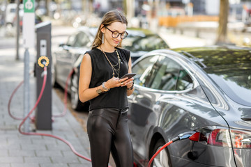 Fototapeta na wymiar Young woman charging electric car standing with smart phone outdoors on the street in Rotterdam city