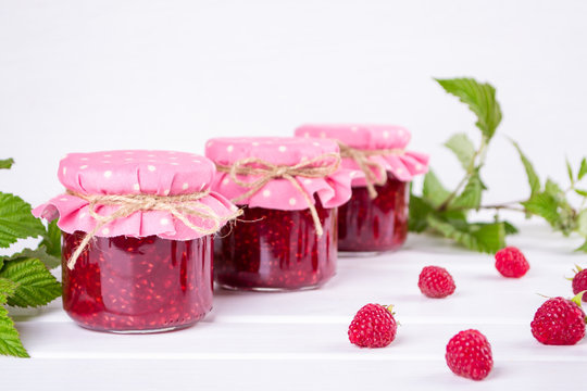 Jars with raspberry jam placed in rows and fresh raspberry on white wooden table