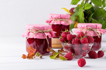 Raspberry jam in glass jar, fresh  ripe raspberry and green leaves on white wooden table