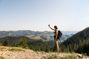 Young female backpacker uses mobile phone for self portrait in rural mountain area of ukrainian carpathian mountains. Girl with tourist rucksack on a long hiking walk looking for network connection