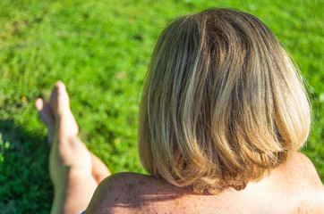 Rear view of woman head. Woman with blond hair and naked shoulders sitting in a green grass meadow.