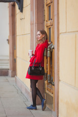 Portrait of a beautiful blonde girl with long hair in a red coat walking along the street. Girl holding coffee in hands
