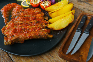 Pork Spare Ribs Barbecue with fried potato and fresh vegetable in black ceramic dish with knife on wooden table