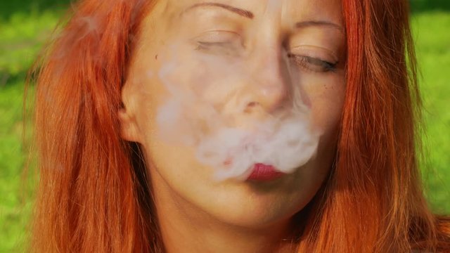 Cute girl with red hair at the green grass park and smokes vape with sunlight close-up