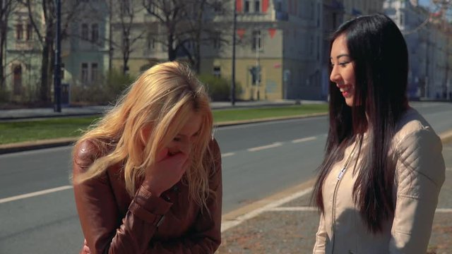 A young Caucasian woman and a young Asian woman talk and laugh together in a street in an urban area