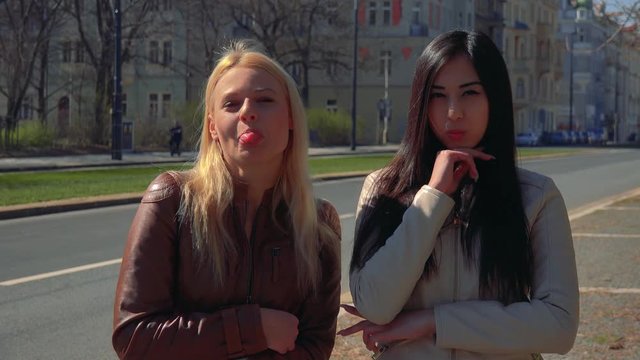 A young Caucasian woman and a young Asian woman make goofy faces and strike goofy poses in a street in an urban area