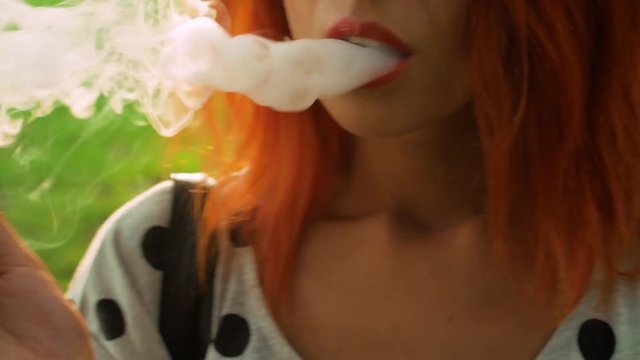Beautiful girl with red hair smokes vape in summer park close-up