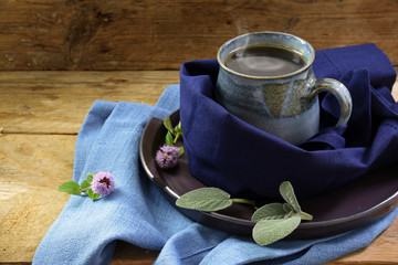 Cup of hot steaming herbal tea against cold wrapped in a blue cloth on rustic wood, fresh herbs as sage and mint  for the immune system and flu prevention, copy space, selected focus