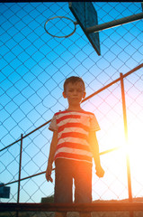 The boy stands behind the net on the basketball court against the backdrop of the control sunset.