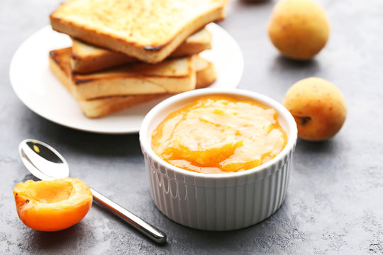 Apricot jam in bowl with bread toasts on grey wooden table