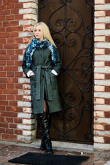 Стильная красивая девушка со светлыми волосами стоит возле железной двери в сером пальто, сапогах и синем шарфе 