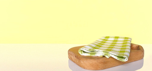 Cutting board with checkered green tablecloth on white table  over yellow wall. Background with copy space.