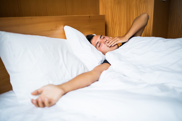 Cheerful young man is waking up after sleeping in the morning. He is yawing His eyes are closed with relaxation.